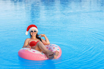 smiling young woman in santa hat and sunglasses relaxing on inflatable ring in swimming pool