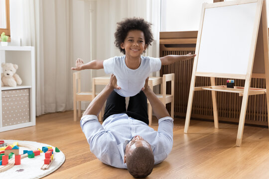 Happy preschool little Black kid playing airplane with strong dad. Father holding son with flying hands in air, lying on warm floor, exercising with child, practicing support, balance, acroyoga