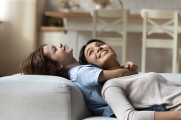 Attractive Asian young mother and little son cuddling lying together on sofa, close up. Loving family relaxing at home, daydreaming enjoy carefree weekend in fashionable flat. Love, parenthood concept