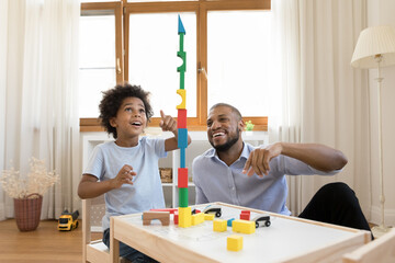 Excited Black little son and happy dad building high toy tower together, playing with construction...