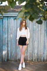 A girl in a white top and valuable shorts on the background of an old fenc