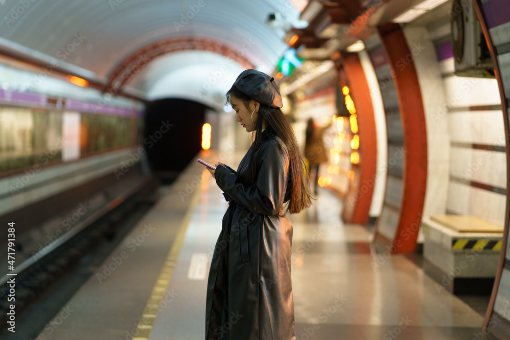 Sticker Korean girl with smartphone at empty metro platform. Stylish asian female browsing social media waiting for underground train. Young woman dependent from mobile phone and online communication concept