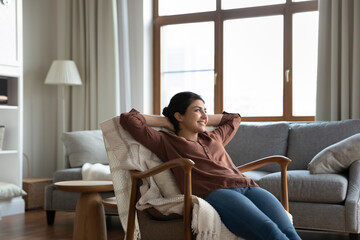 Obraz premium Carefree happy millennial indian woman resting in cozy armchair with folded arms behind head, enjoying peaceful calm stress free leisure weekend time alone in modern living room, breathing fresh air.