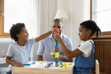 Happy Black kids playing together building high plastic tower on desk for board game, completing model from toy construction blocks. Dad watching little sibling children at home. Childhood, daycare