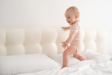 Cute baby girl in bodysuit stand holding bed frame, learning to walk on white bedding on bed