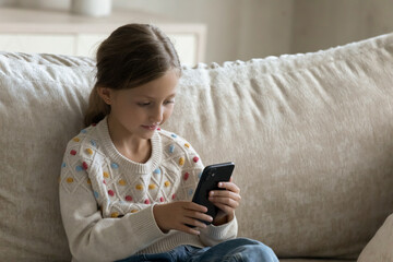 Happy focused little Z generation cute kid girl using cellphone, sitting alone on comfortable couch, playing entertaining mobile games, watching funny videos in social networks, spending time online.