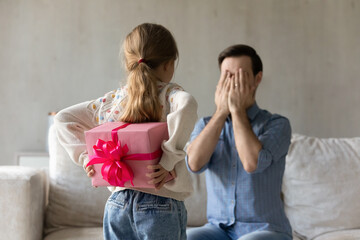 Rear view happy small child daughter holding wrapped gift box behind back, looking at daddy...