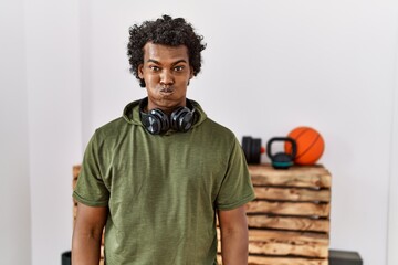 African man with curly hair wearing sportswear at the gym puffing cheeks with funny face. mouth...