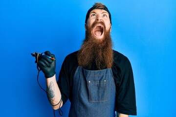 Redhead man with long beard tattoo artist wearing professional uniform and gloves angry and mad...