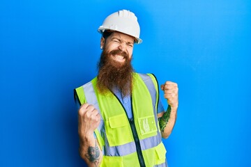 Redhead man with long beard wearing safety helmet and reflective jacket very happy and excited doing winner gesture with arms raised, smiling and screaming for success. celebration concept.