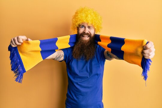 Redhead man with long beard football hooligan cheering game wearing funny wig winking looking at the camera with sexy expression, cheerful and happy face.