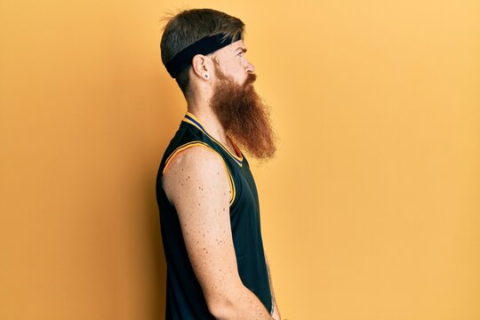 Redhead man with long beard wearing basketball uniform looking to side, relax profile pose with natural face with confident smile.