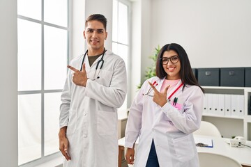 Young doctors wearing uniform and stethoscope at the clinic smiling cheerful pointing with hand and finger up to the side