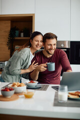 Happy couple surfing the net on laptop while having breakfast at dining table.