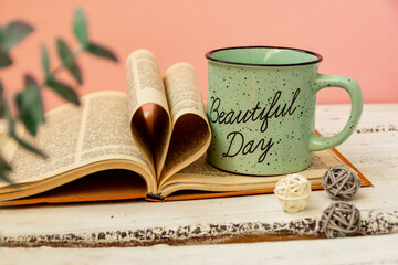 A green cup of tea stands on a book. Pink background. Eucalyptus flowers. The pages of the book are wrapped in the shape of a heart