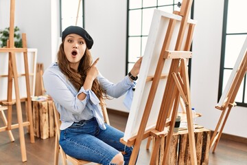 Young hispanic artist woman painting on canvas at art studio surprised pointing with finger to the side, open mouth amazed expression.