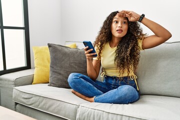Beautiful hispanic woman sitting on the sofa at home using smartphone very happy and smiling looking far away with hand over head. searching concept.