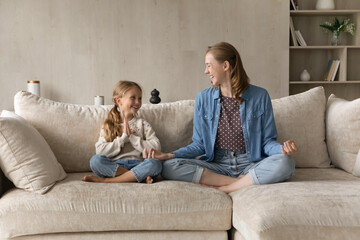 Obraz premium Cheerful little 6s kid girl sitting in lotus position with caring young mother on comfortable couch, learning doing yoga calming breathing exercises together in modern living room, hobby activity.
