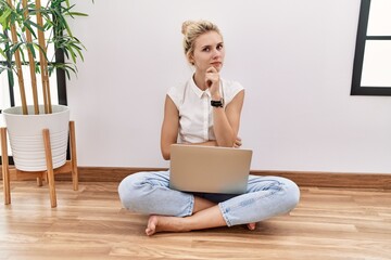 Young blonde woman using computer laptop sitting on the floor at the living room looking confident...