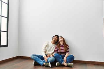 Young hispanic couple looking to the ceiling sitting on the floor at empty new home.