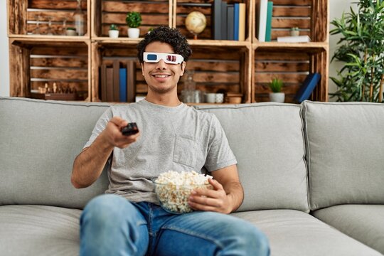 Young hispanic man watching movie using 3d glasses sitting on the sofa at home.