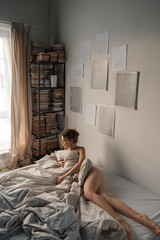 Young beautiful blond woman lies in bed. A naked woman covers herself with a blanket. Sunny morning...