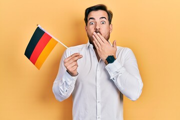 Handsome caucasian man with beard holding germany flag covering mouth with hand, shocked and afraid...