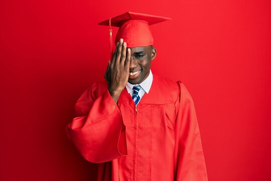 Young african american man wearing graduation cap and ceremony robe covering one eye with hand, confident smile on face and surprise emotion.