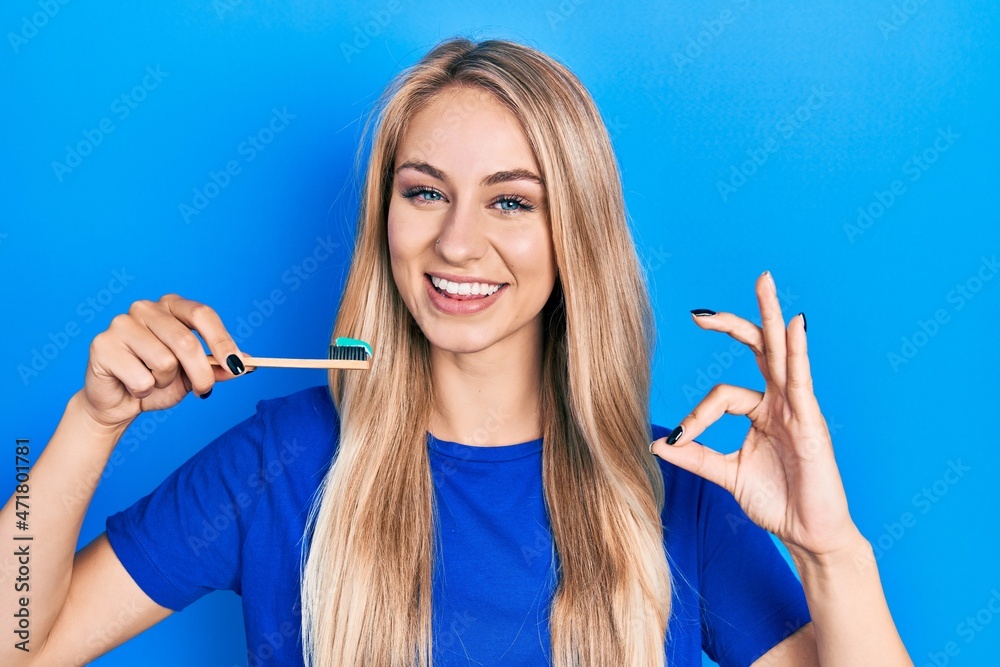 Sticker young beautiful caucasian woman holding toothbrush with toothpaste doing ok sign with fingers, smili