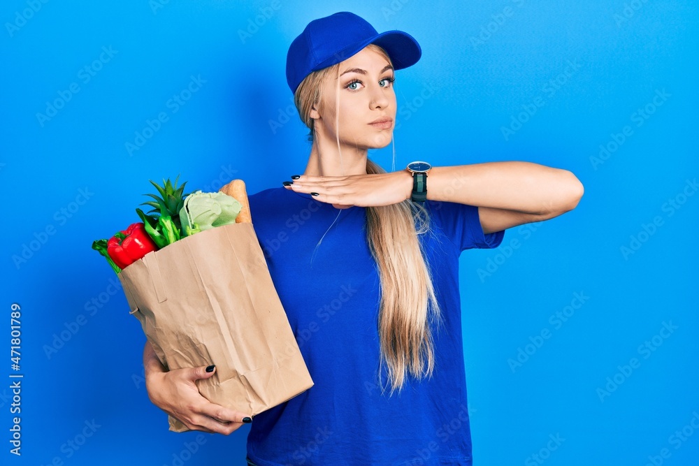 Poster Young caucasian woman wearing courier uniform with groceries from supermarket cutting throat with hand as knife, threaten aggression with furious violence