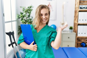 Young caucasian woman working at pain recovery clinic smiling cheerful presenting and pointing with palm of hand looking at the camera.