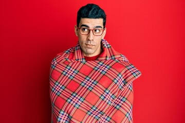 Handsome hispanic man wrapped in a red warm red blanket smiling looking to the side and staring away thinking.