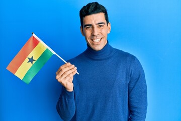 Handsome hispanic man holding ghana flag looking positive and happy standing and smiling with a confident smile showing teeth