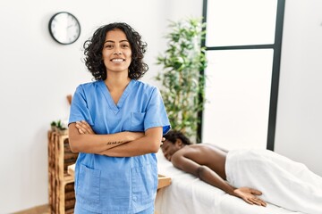 Young physiotherapist woman smiling happy standing with arms crossed gesture. at the clinic. African american man waiting to recive massage at the clinic. - obrazy, fototapety, plakaty