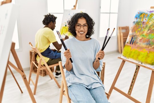 African american artist couple smiling happy painting at art studio. Woman holding paintbrushes sitting on chair.