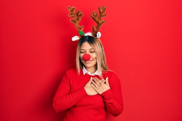 Beautiful hispanic woman wearing deer christmas hat and red nose smiling with hands on chest with closed eyes and grateful gesture on face. health concept.