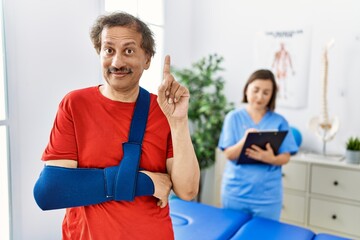 Fototapeta premium Southeast asian man wearing wearing arm on sling at rehabilitation clinic surprised with an idea or question pointing finger with happy face, number one
