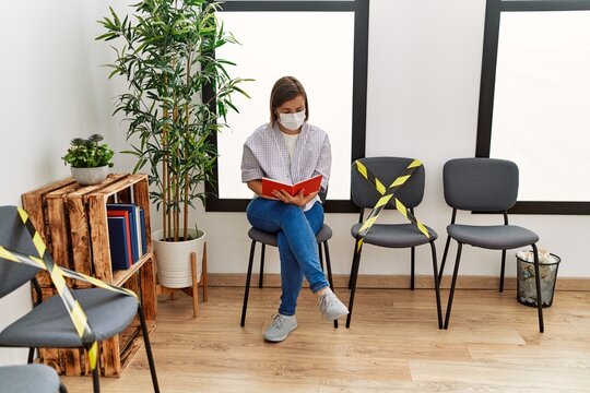 Middle Age Hispanic Woman Wearing Safety Mask Reading A Book At Waiting Room