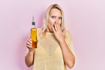 Beautiful caucasian blonde woman holding olive oil bottle covering mouth with hand, shocked and...