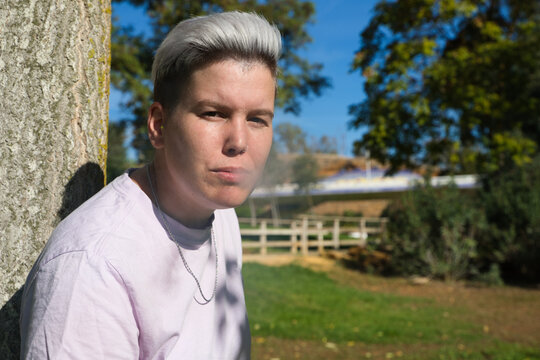 gender non-binary and androgynous person is leaning against a tree lighting a cigarette and smoking. He is looking at the camera. Concept of non-binary and androgynous. Tobacco is harmful to health