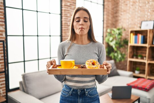 Young woman holding tray with breakfast food making fish face with mouth and squinting eyes, crazy and comical.