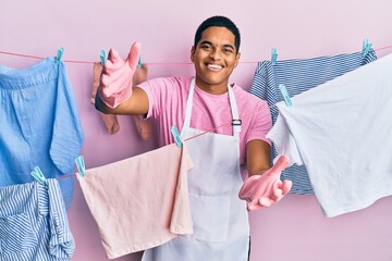 Young handsome hispanic man wearing cleaner apron holding clothes on clothesline looking at the...