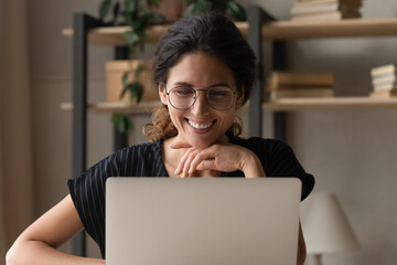 Good work. Positive smiling latina woman wear glasses sit by desk use laptop engaged in web search of information write blog online. Happy young female chat at internet read good news at social media - obrazy, fototapety, plakaty