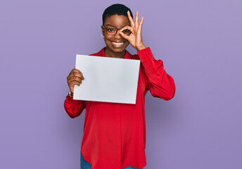 Young african american woman holding blank empty banner smiling happy doing ok sign with hand on eye looking through fingers