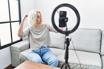 Young caucasian woman recording vlog tutorial with smartphone at home showing arms muscles smiling proud. fitness concept.