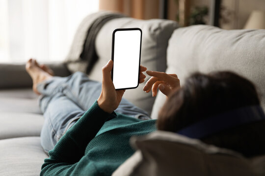 Female Using Phone. Over Shoulder View Of Young Woman Lying On Sofa Hold Smartphone With Blank Empty Screen. Template For Web App Chat Interface Online Advertisement Mobile Game Social Network Profile