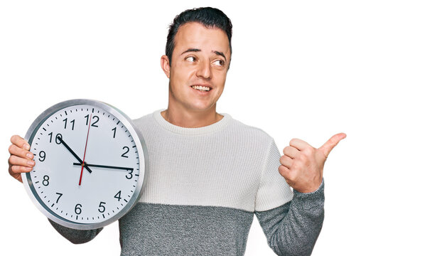 Handsome young man holding big clock pointing thumb up to the side smiling happy with open mouth
