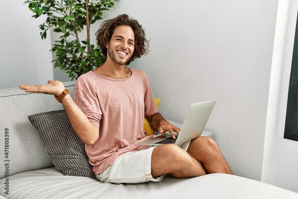 Sticker young hispanic man sitting on the sofa at home using laptop smiling cheerful presenting and pointing