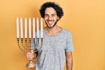 Handsome hispanic man holding menorah hanukkah jewish candle looking positive and happy standing...