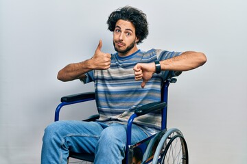 Handsome hispanic man sitting on wheelchair doing thumbs up and down, disagreement and agreement expression. crazy conflict